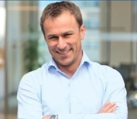 Ein lächelnder Mann mit kurzen, braunen Haaren steht in einem modernen Büro und hat die Arme vor der Brust verschränkt.